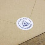 Kleiner runder Aufkleber mit Segelschiff und Text "Mast- und Schotbruch" auf braunem Papier.