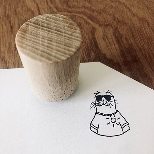 Hölzerner Stempel auf weißem Papier mit Katzenzeichnung in Sonnenbrille und T-Shirt.