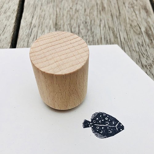 Ein Holzstempel auf weißem Papier mit Fischzeichnung, auf einem Holztisch.