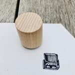 Rundes Holzstück auf weißem Papier mit Stempelabdruck, natürliche Holzmaserung sichtbar.