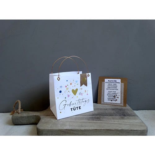 Geburtstagsgeschenk-Tüte mit bunten Punkten und Herz, auf Holzplatte vor grauer Wand.