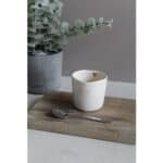 Hochwertige weiße Tasse mit Herzdekor auf Holztablett, Eukalyptus im Hintergrund, stilvolle Tischdek.