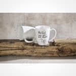 Ein weißer Kaffeebecher mit handgeschriebenem Text und Zeichnung, auf Holzstamm liegend, rustikaler.