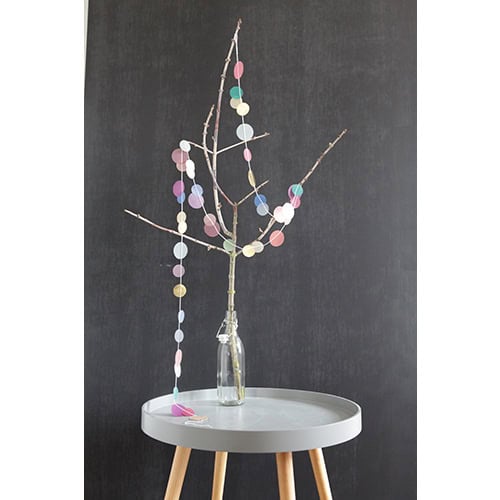 Blumenzweig mit bunten Papierkreisen in einer Glasflasche auf einem weißen Tisch.