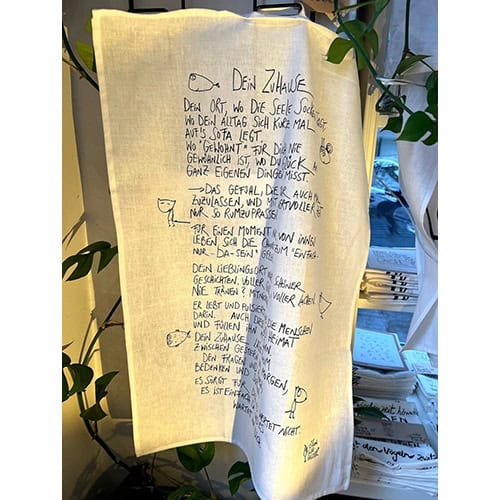 Handgeschriebener Text auf einem weißen Stoff, umgeben von grünen Pflanzen, im Innenraum.