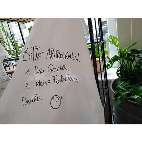 Handgeschriebene Notiz auf Papier mit Bitte um Abtrocknen, im Café mit Pflanzen im Hintergrund.