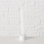 Weiße Kerze in Kerzenhalter vor weißer Ziegelwand, minimalistisch und elegant.