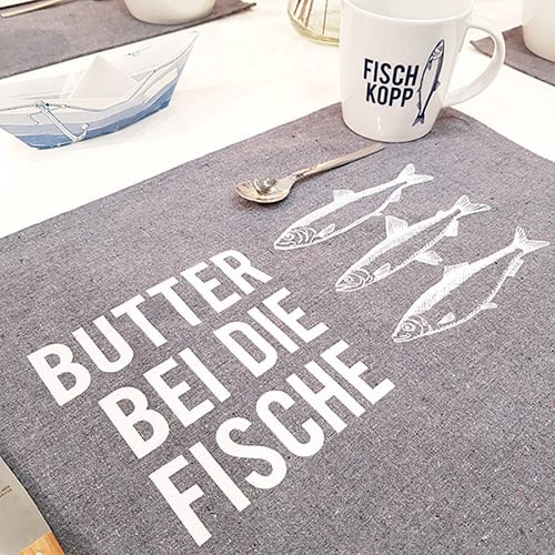 Dekorativer Tischläufer mit Fischmotiven und Spruch "Butter bei die Fische" in modernem Design.