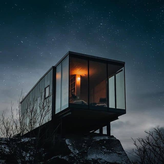 Modernes Haus auf Felsen bei Nacht mit Sternenhimmel, Glasfassade, warmem Licht.