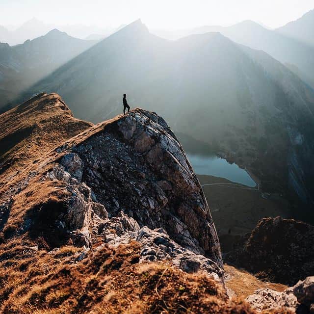 Bergsteiger steht auf einem Felsen mit Blick auf die umliegenden Berge und einen See in den Alpen.