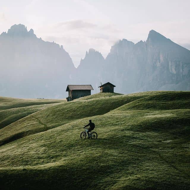 Radfahrer auf grünen Hügeln vor Bergen und Hütten in der Natur.