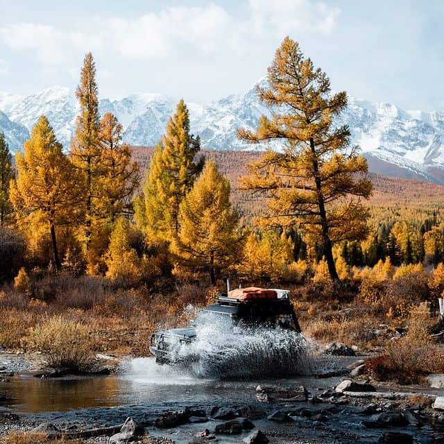 Geländewagen fährt durch Fluss inmitten von Herbstbäumen und Bergen.