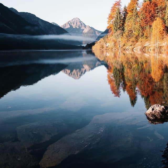 Bergsee mit ruhigem Wasser, umgeben von bunten Herbstbäumen und hohen Bergen im Hintergrund.