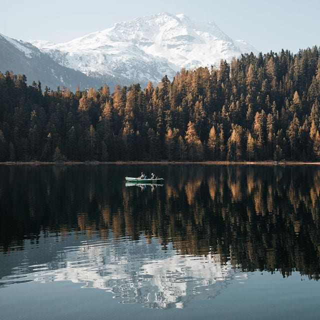 Malerischer Bergsee mit Boot, umgeben von Tannen und schneebedecktem Gipfel im Hintergrund.