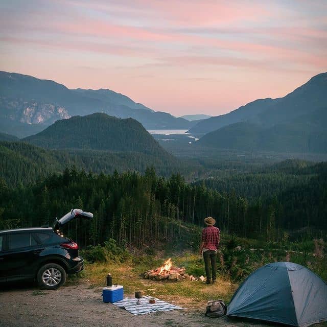Natur- und Campinglandschaft in den Bergen mit Zelt, Auto und Lagerfeuer bei Sonnenuntergang.