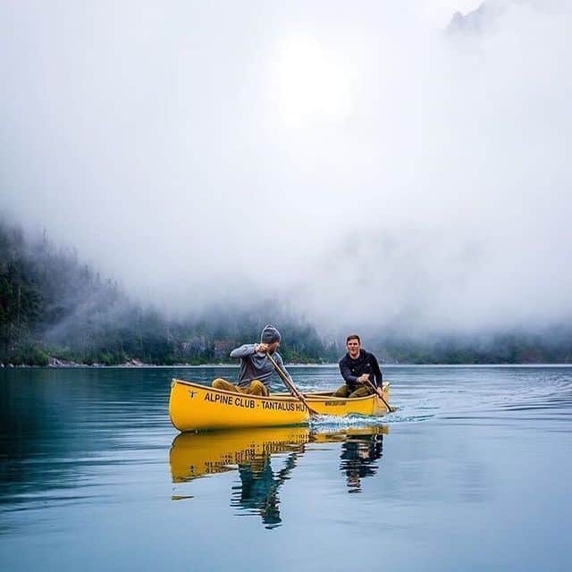 Zwei Personen paddeln in einem gelben Kanu auf einem stillen Bergsee, umgeben von Nebel und Bergen.