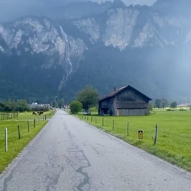 Ländliche Straße führt zu einem alten Holzhaus vor beeindruckender Bergkulisse mit Wasserfall im Hin.