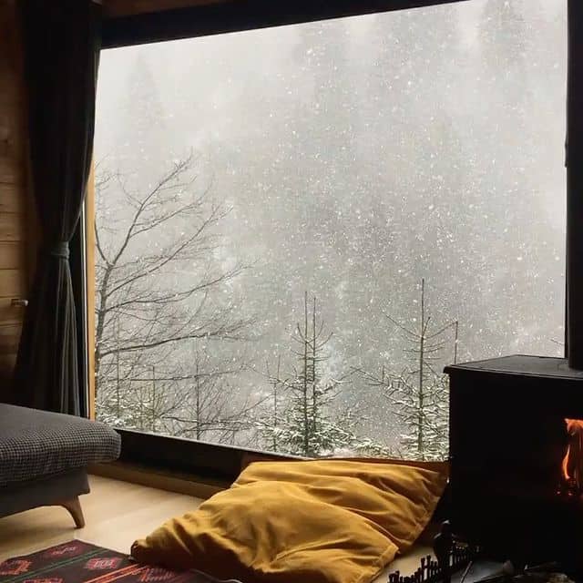 Gemütliches Wohnzimmer mit Blick auf verschneite Bäume durch große Fenster, Kamin und bequeme Sitzge.