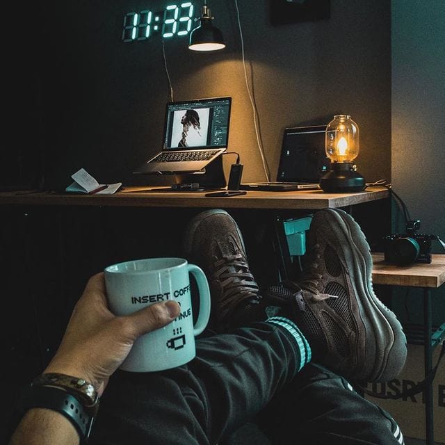 Gemütlicher Arbeitsplatz mit Laptop, Kaffee und Uhr im Hintergrund.