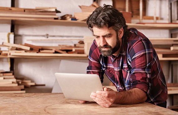 Mann mit Bart und kariertem Hemd arbeitet mit Tablet in Holzwerkstatt.