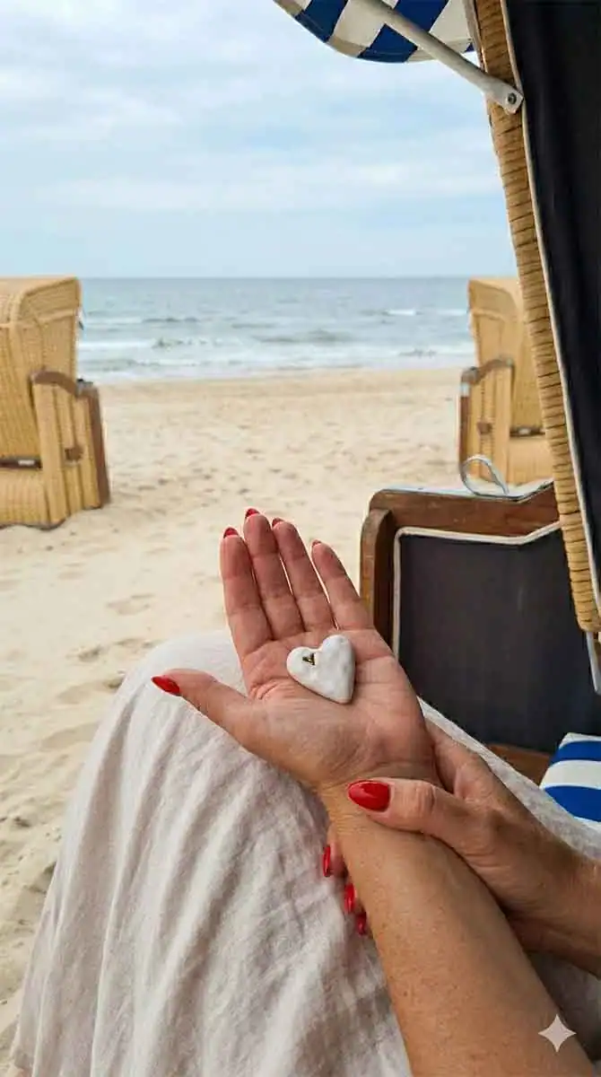 Frau hält Herzstein am Strand, Blick aufs Meer, entspannte Atmosphäre.