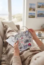 Person hält handgezeichnete Leuchtturm- und Hafenbilder, entspannt im Wohnzimmer.